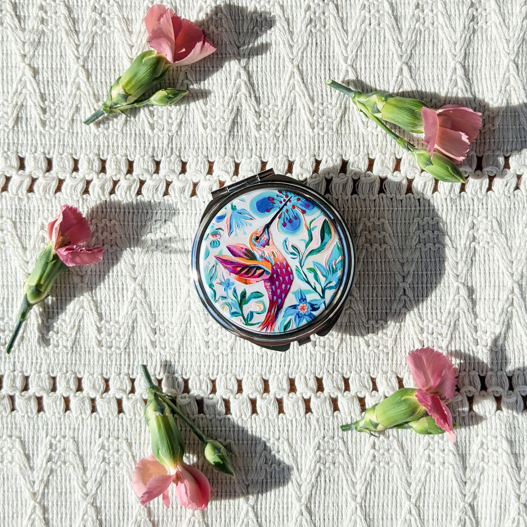 Round mirror with colorful hummingbird design on a textured surface with pink flowers