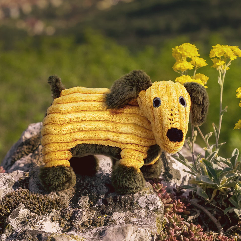 Yellow dog-shaped plush toy on a rock with greenery and flowers in the background