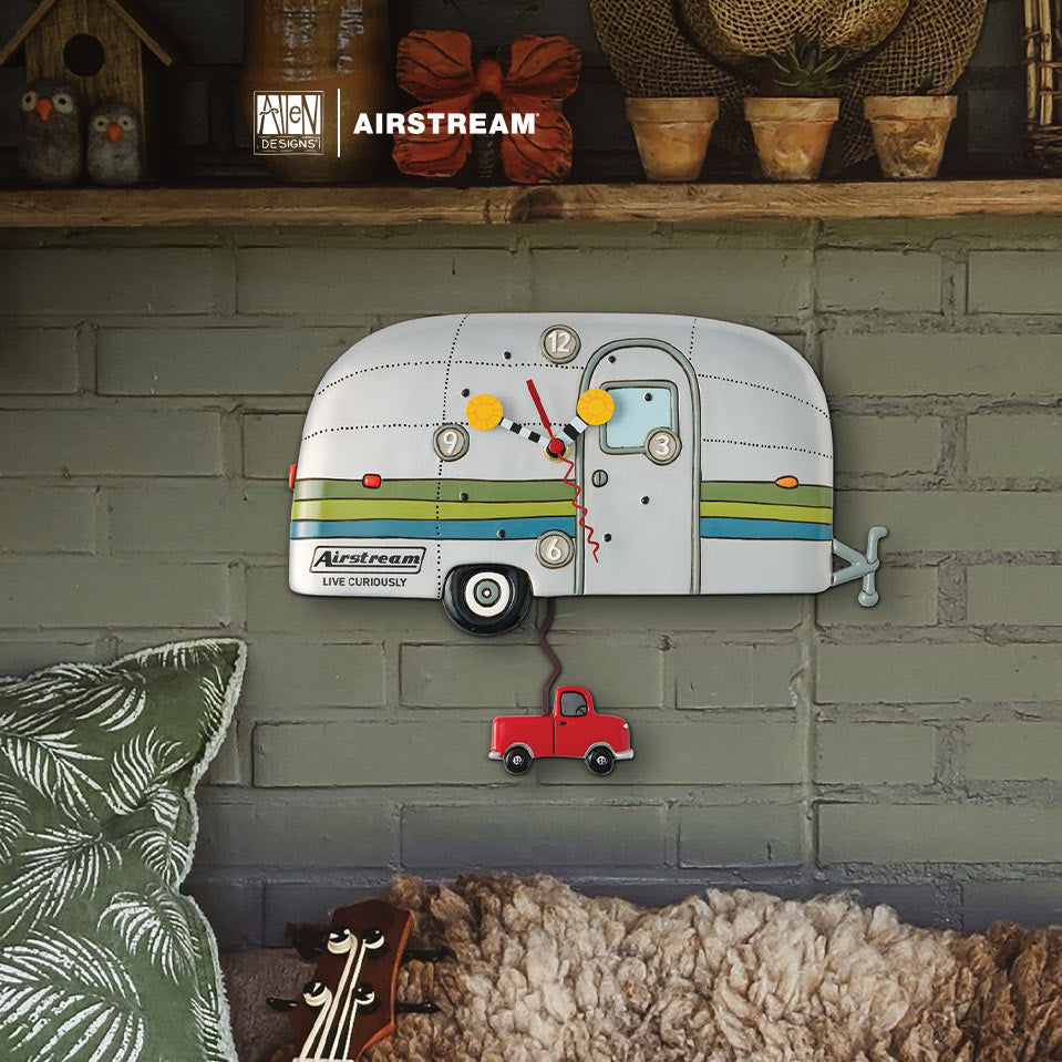 A vintage-style trailer wall clock with a red hanging truck and colorful stripe details, mounted on a painted brick wall above a bed with a pillow and blanket. The Airstream logo is visible on the clock and in the top banner.