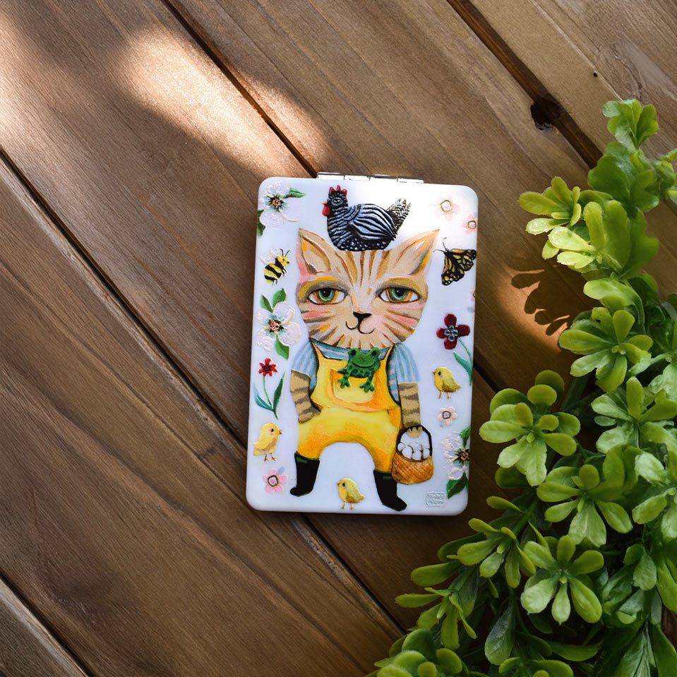 A rectangular metal tin with a whimsical illustration of a cat dressed as a farmer, surrounded by small birds, bees, flowers, and chicks. The tin is placed on a wooden deck next to a green plant.