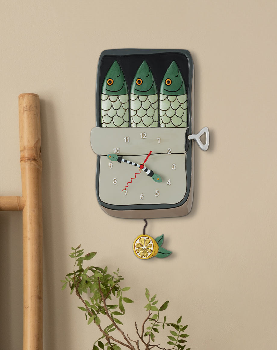 A whimsical wall clock shaped like a fisherman's scale with three green fish on top, a gray dial, and a hanging lemon charm, mounted on a beige wall next to a bamboo pole.