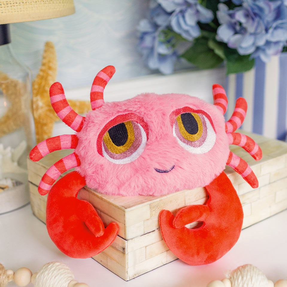 A pink plush crab toy with large cartoon eyes, red and pink striped antennae, and bright red claws, displayed on a small wooden box in a styled indoor setting with flowers and seashells.