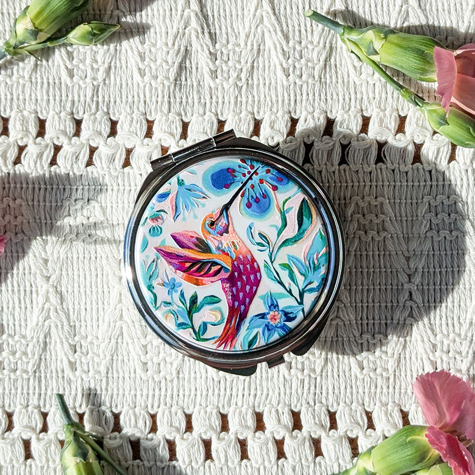 Compact mirror with colorful bird design on a textured white surface with flowers.