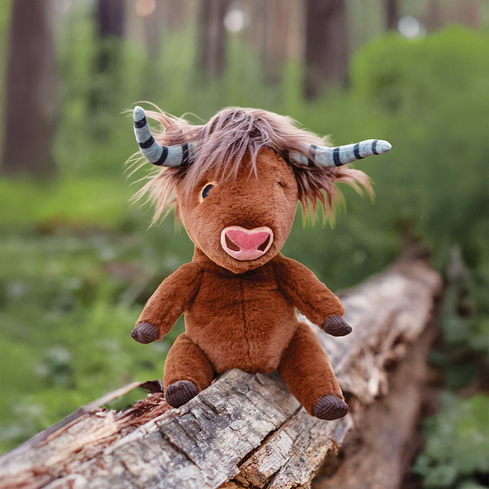 A brown plush Highland cow toy with shaggy pinkish-brown hair, striped horns, and a pink nose, sitting on a fallen log in a blurred forest setting.
