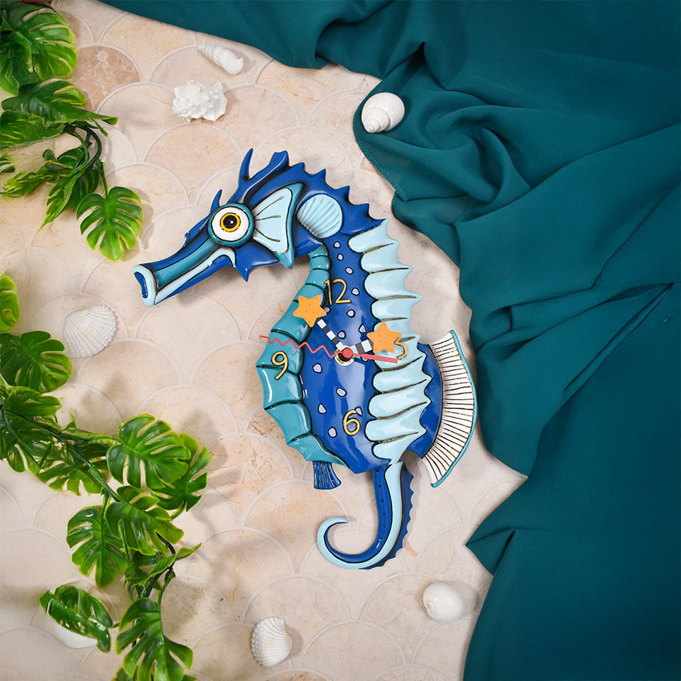 A decorative blue seahorse wall clock styled in a flat-lay arrangement with green artificial leaves, white seashells, and draped teal fabric on a beige patterned surface.