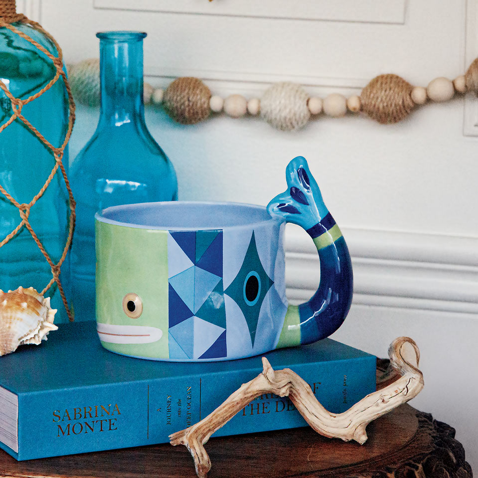 A ceramic mug shaped like a fish, featuring a light green face and a geometric blue pattern, set on a blue book with driftwood and a turquoise glass bottle in the background.